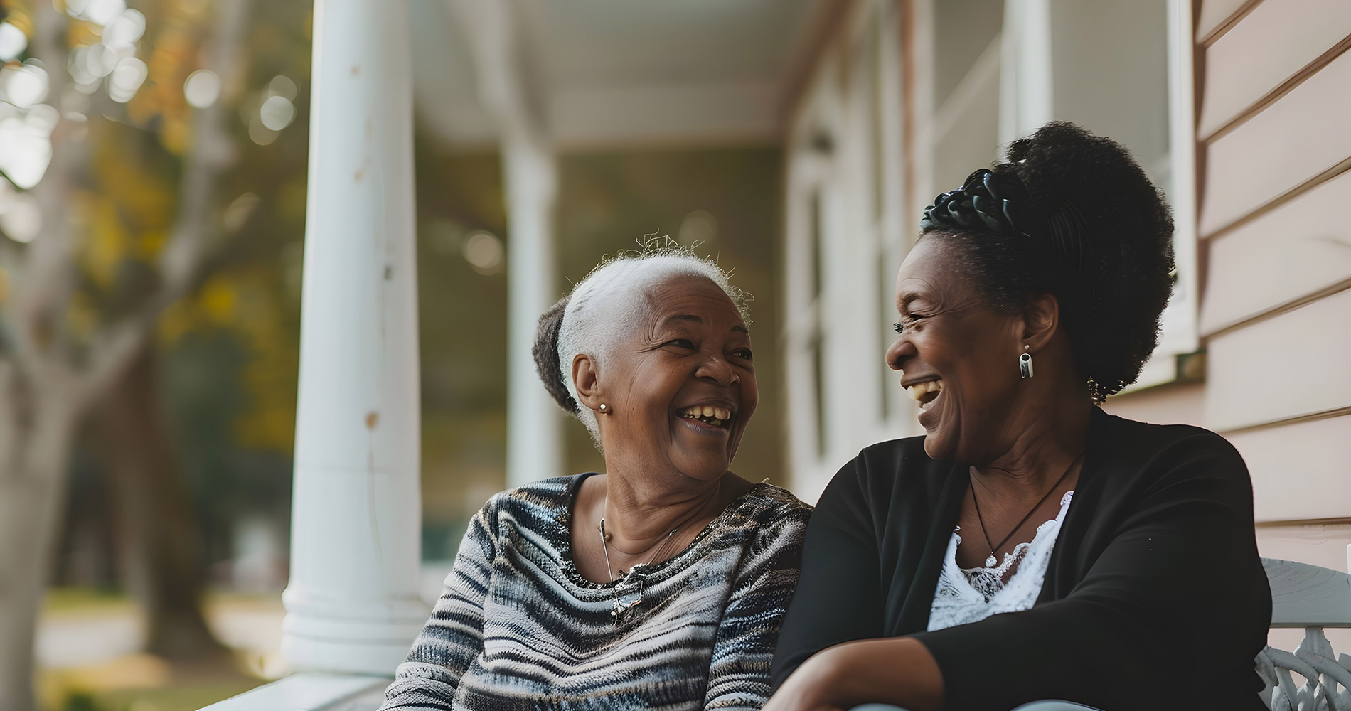 Two older women sit on a porch bench, facing each other and smiling, with trees and columns in the background.