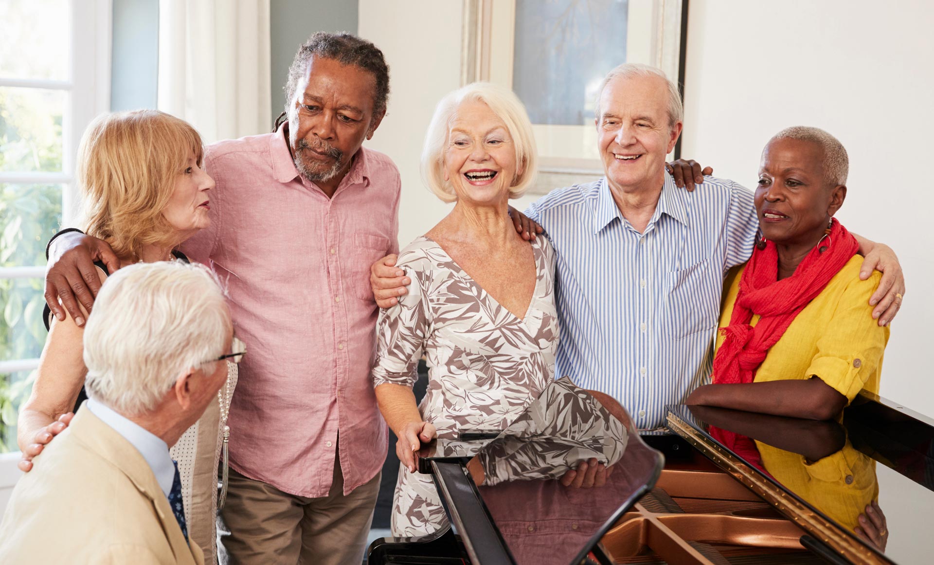Six older adults stand and sit together around a piano indoors, smiling and talking in a well-lit room.